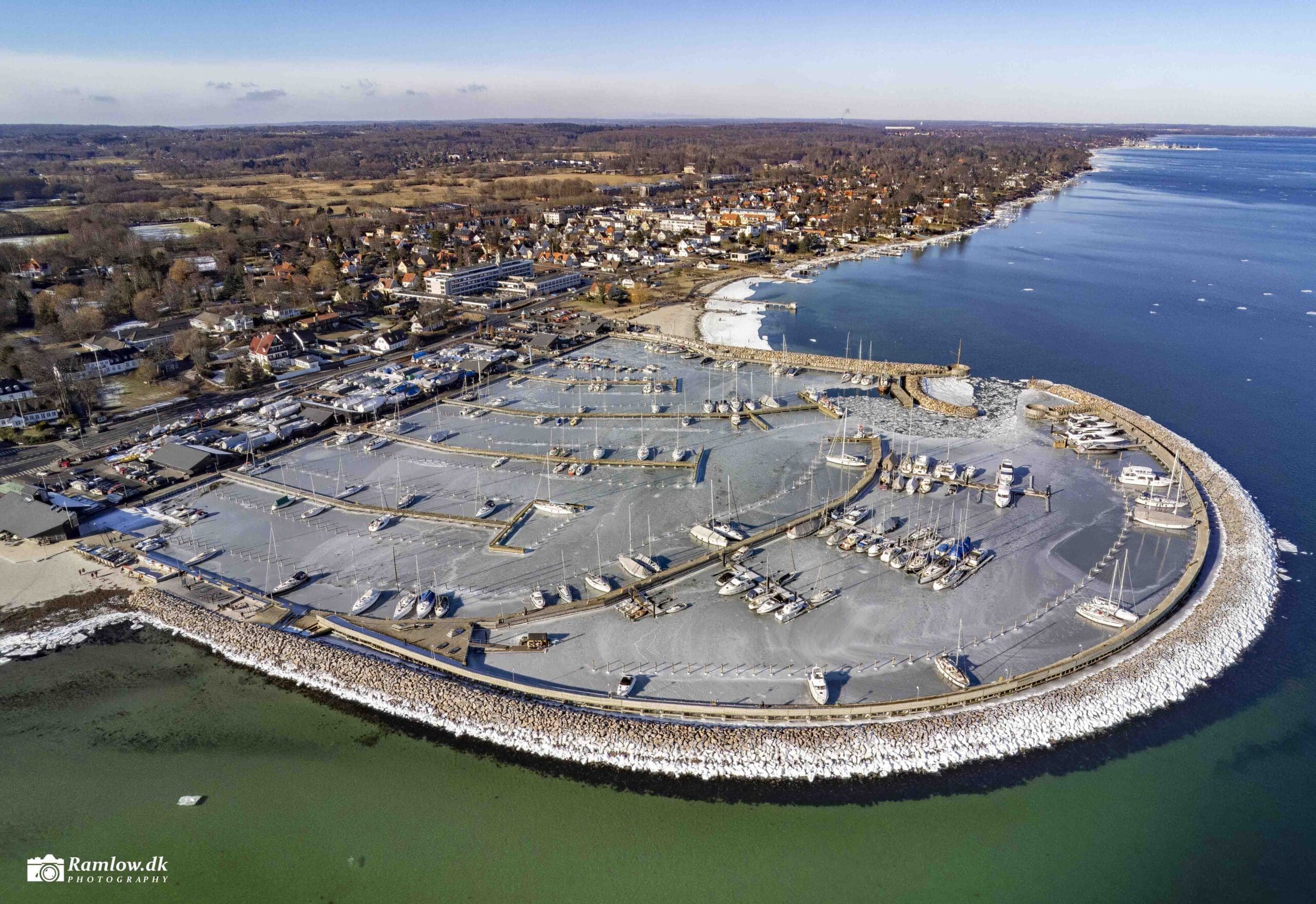 Drone-skud – Vedbæk Havn i vinterklæder med islagt havnebassin, bådebroer og Vedbæk i baggrunden
